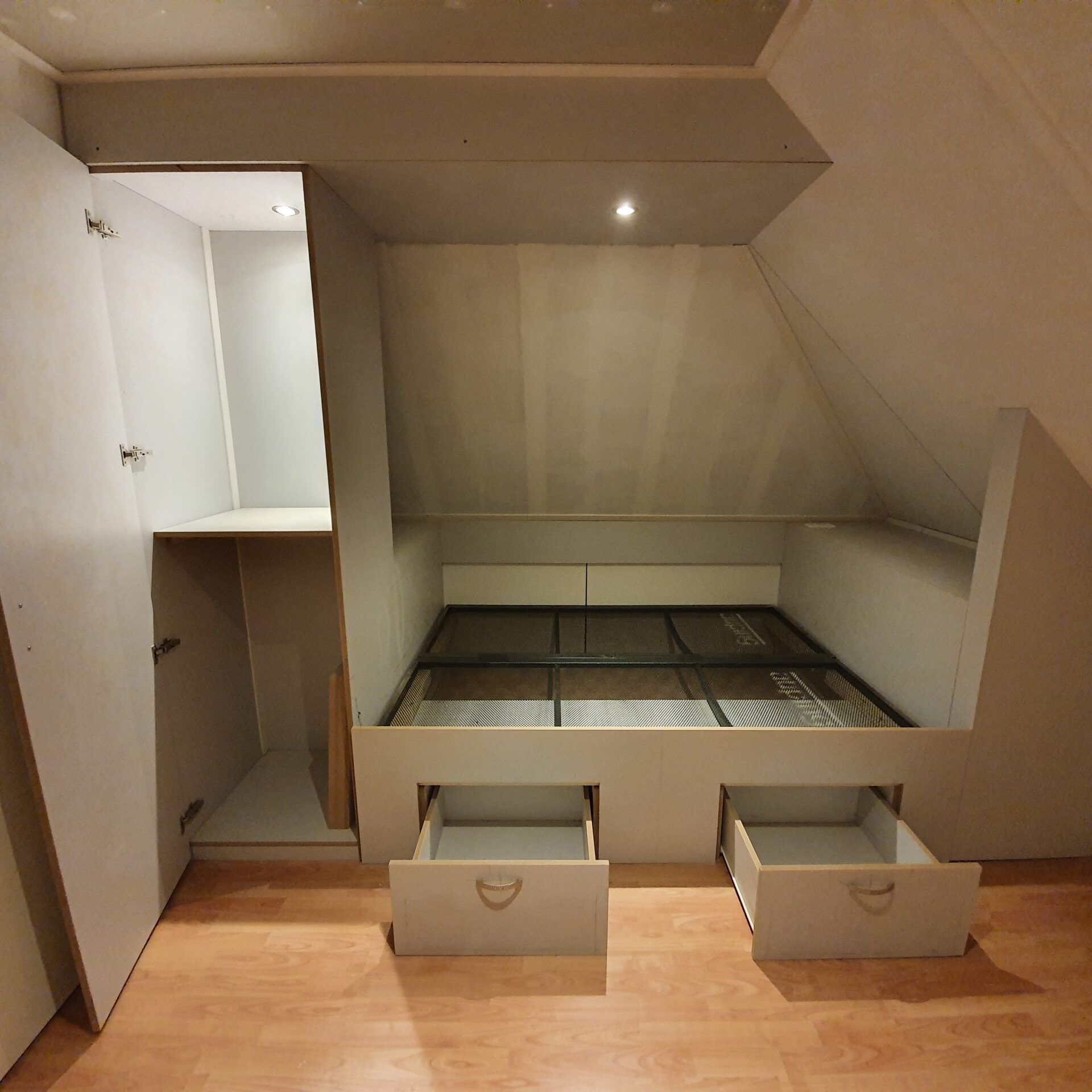 Ingebouwd bed met open lades en kast in een zolderruimte met schuine wanden en ingebouwde verlichting