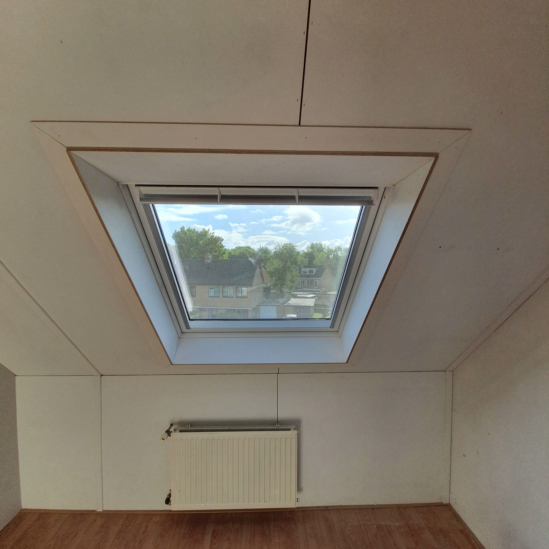 Zolderruimte met een dakraam met rolluik, witte radiator en houten vloer